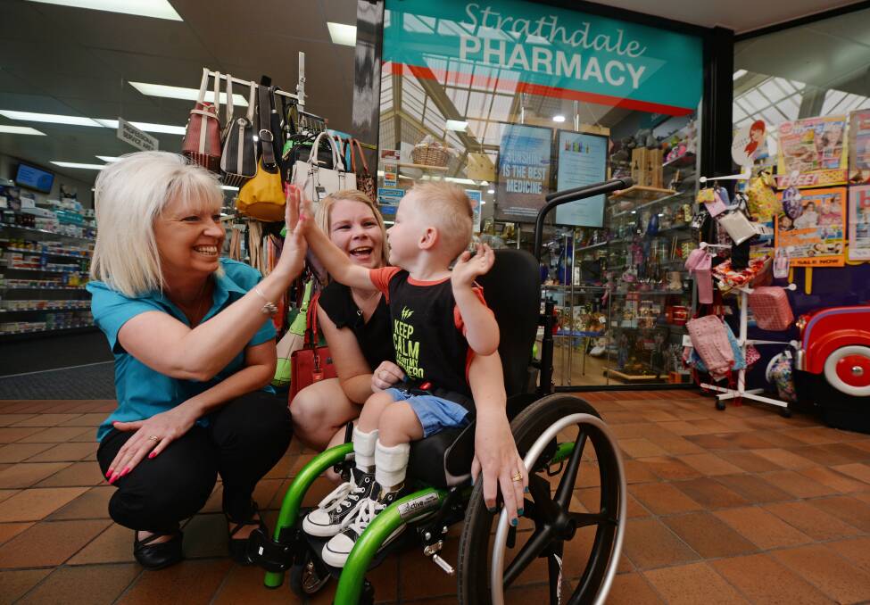 Jett McNamara high fives Strathdale Pharmacy's Caroline Marsh in his new wheelchair while his mother Amy looks on. Picture: DARREN HOWE